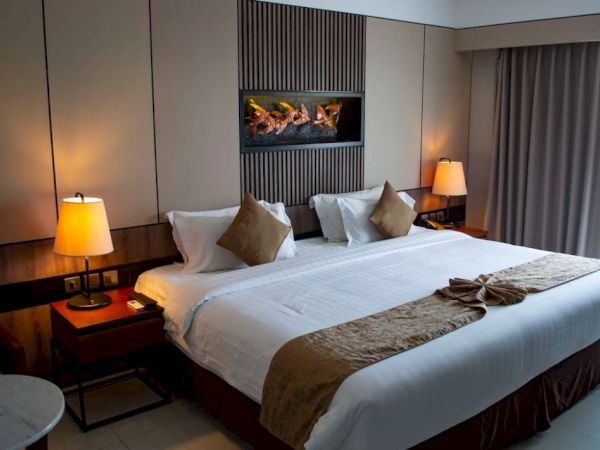 A cozy hotel room with a large bed, pillows, side tables with lamps, and a decorative fireplace on the wall.