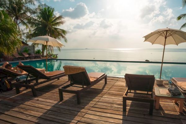 A serene poolside scene with lounge chairs, umbrellas, and palm trees, overlooking a calm body of water and a bright, cloudy sky.