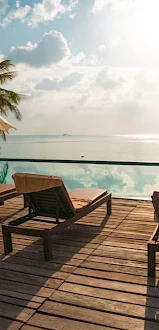 A tropical poolside scene with lounge chairs, umbrellas, and palm trees, overlooking the ocean under a partly cloudy sky.
