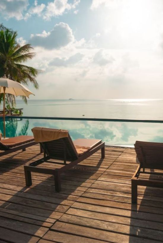Poolside scene with lounge chairs, umbrellas, and palm trees overlooking a tranquil ocean under a partly cloudy sky.