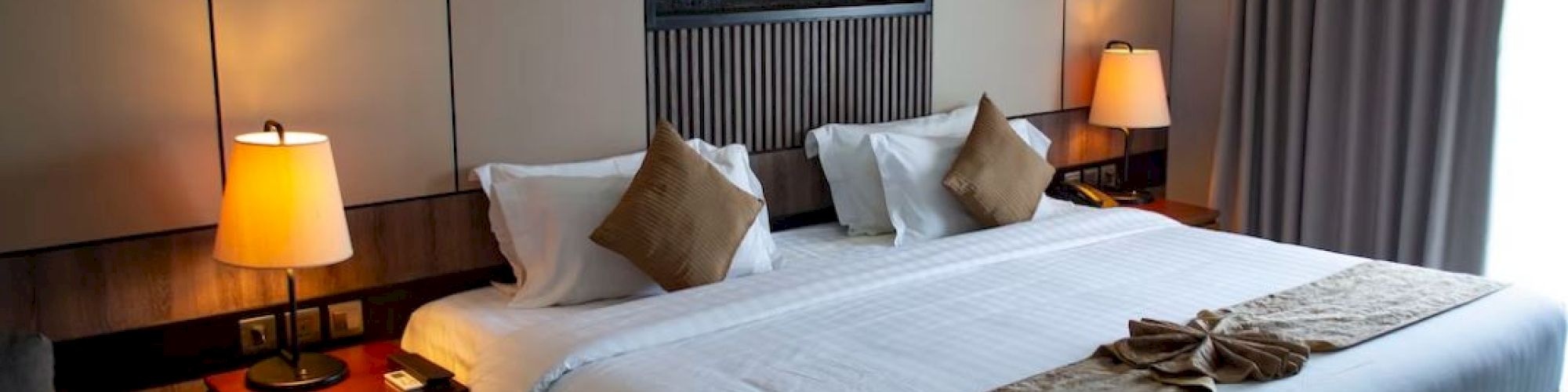 A neatly arranged hotel room with a large bed, cushions, lamps, and a wall-mounted decoration, exuding a cozy and inviting atmosphere.