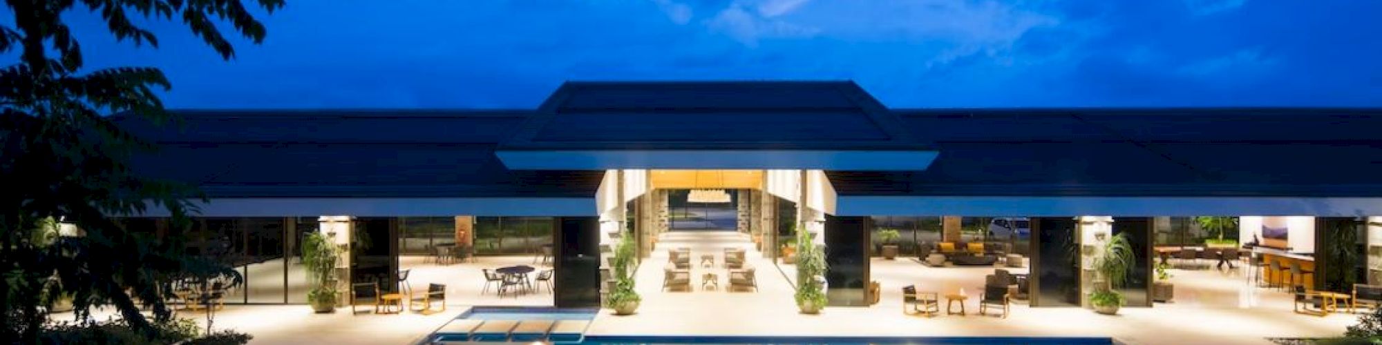 A modern building with outdoor seating is beautifully lit at twilight, featuring a large illuminated pool in the foreground.