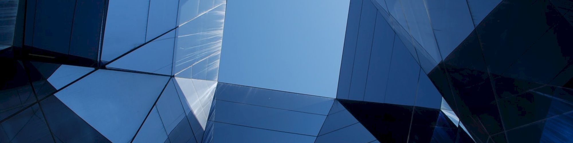 Futuristic architecture with reflective glass surfaces, forming angular shapes and patterns, under a blue sky peeking through at the center.
