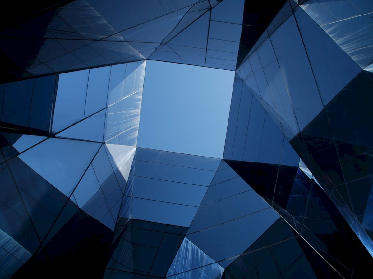 The image shows modern glass architecture forming geometric patterns, with a clear blue sky visible through the structure.