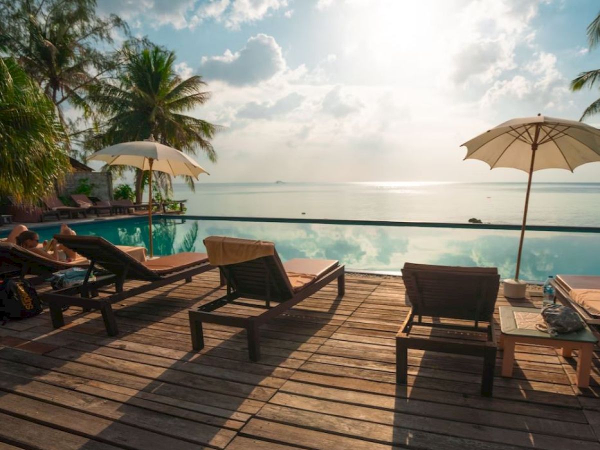 A serene poolside deck with lounge chairs, umbrellas, palm trees, and a tranquil sea view under a partly cloudy sky.