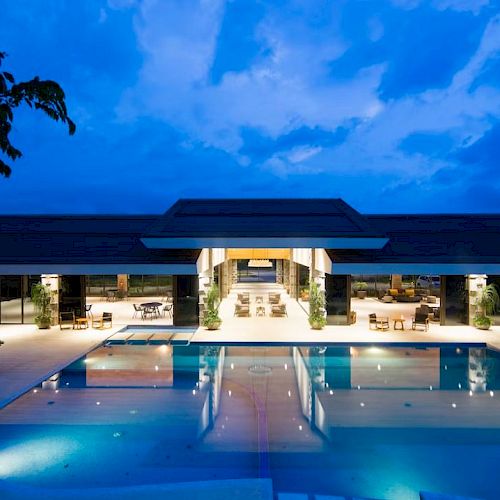 The image shows a luxurious, modern building with a large pool in front, illuminated at dusk, surrounded by some trees.