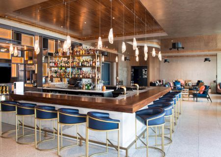 A modern bar with a wooden countertop, blue barstools, overhead lighting, and a well-stocked liquor shelf in a spacious lounge setting.