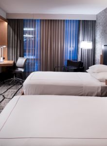 The image shows a modern hotel room with two beds, a flat-screen TV, a desk, and large windows with curtains.