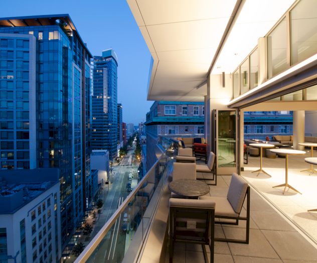A modern rooftop terrace with seating overlooks a cityscape of tall buildings at dusk, with a mix of artificial and natural lighting.