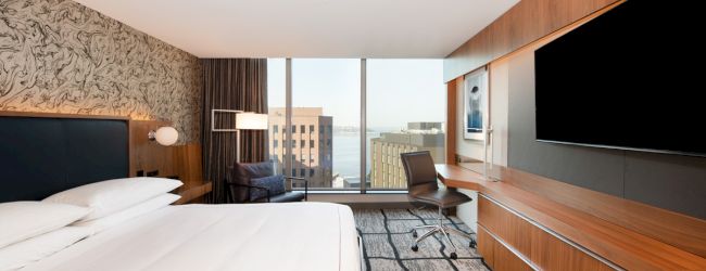 A modern hotel room featuring a large bed, flat-screen TV, desk, chair, and window with a cityscape view, decorated with contemporary style.