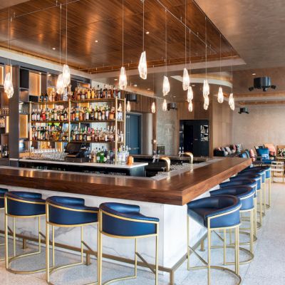 The image shows a modern bar with a central counter, stylish stools, hanging lights, and a well-stocked back shelf.