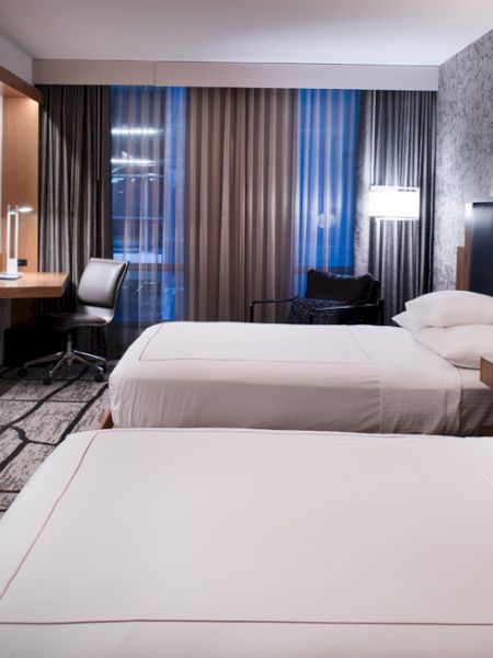The image shows a modern hotel room with two beds, a flat-screen TV, desk, chair, and stylish decor, creating a cozy ambiance.