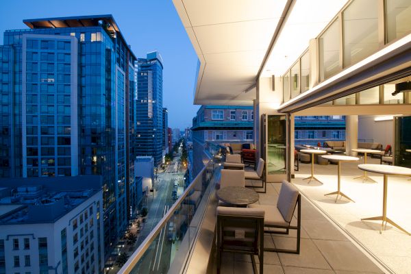A modern cityscape with a view from a balcony, featuring outdoor seating and a night-lit street flanked by tall buildings.