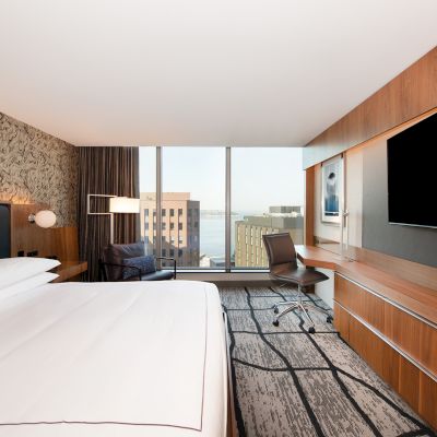 A modern hotel room with a bed, desk, chair, and large TV. Floor-to-ceiling window reveals an urban view with buildings and some water.