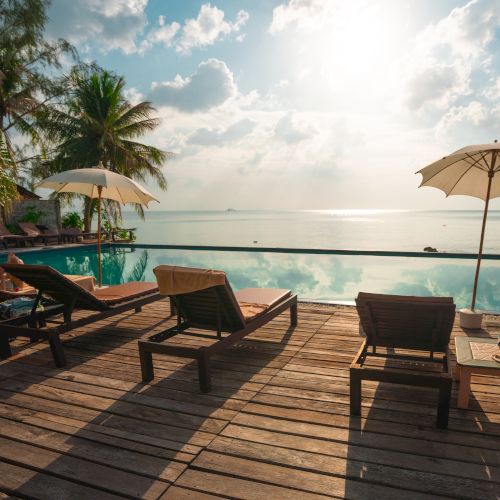 A serene poolside with lounge chairs, umbrellas, palm trees, and a view of the calm sea under a bright sky.