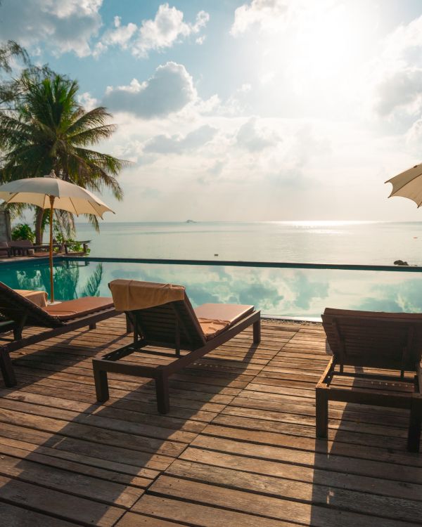 A serene poolside with lounge chairs, umbrellas, palm trees, and a view of the calm sea under a bright sky.