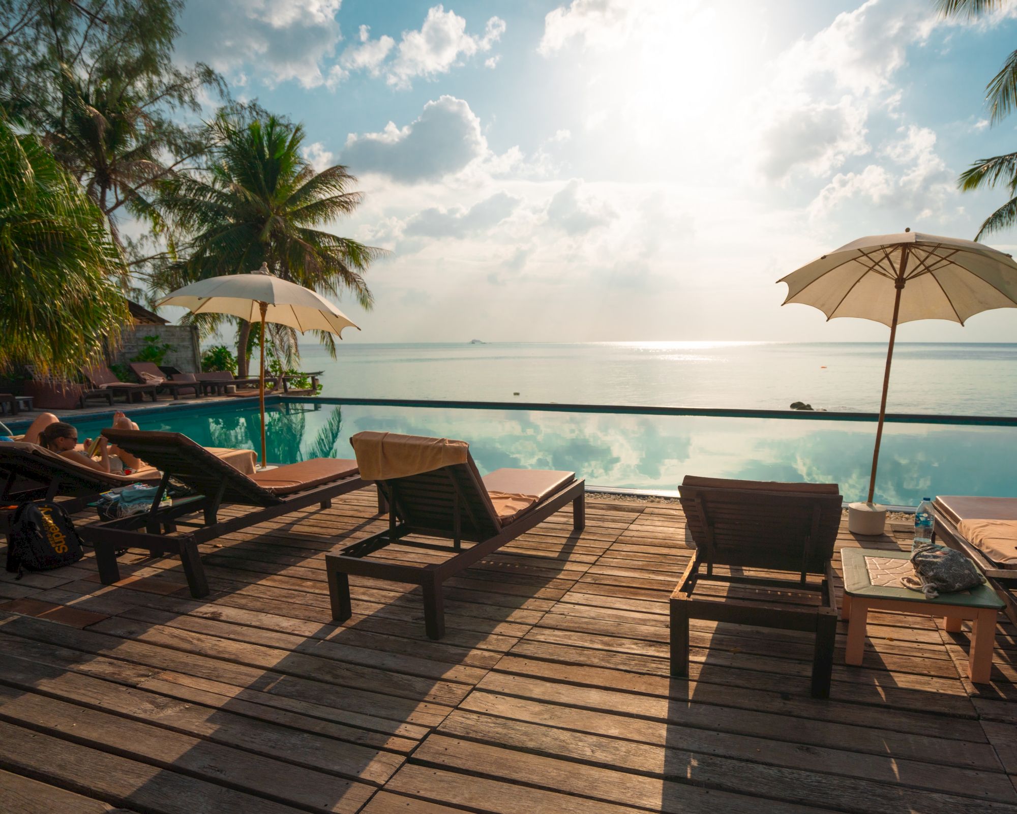 A serene poolside scene with lounge chairs, umbrellas, palm trees, and a view of the ocean under a partly cloudy sky.
