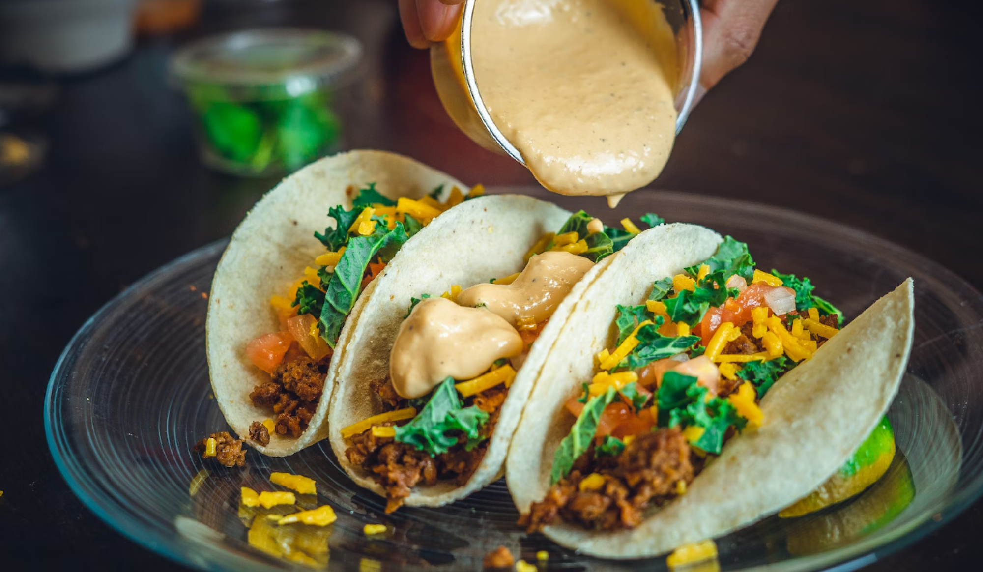 Three tacos filled with ground meat, shredded cheese, kale, tomatoes, and topped with a creamy sauce are served on a glass plate.