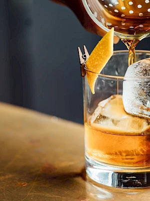 A cocktail being poured into a glass with ice and garnished with a twist of citrus peel on a bar counter.