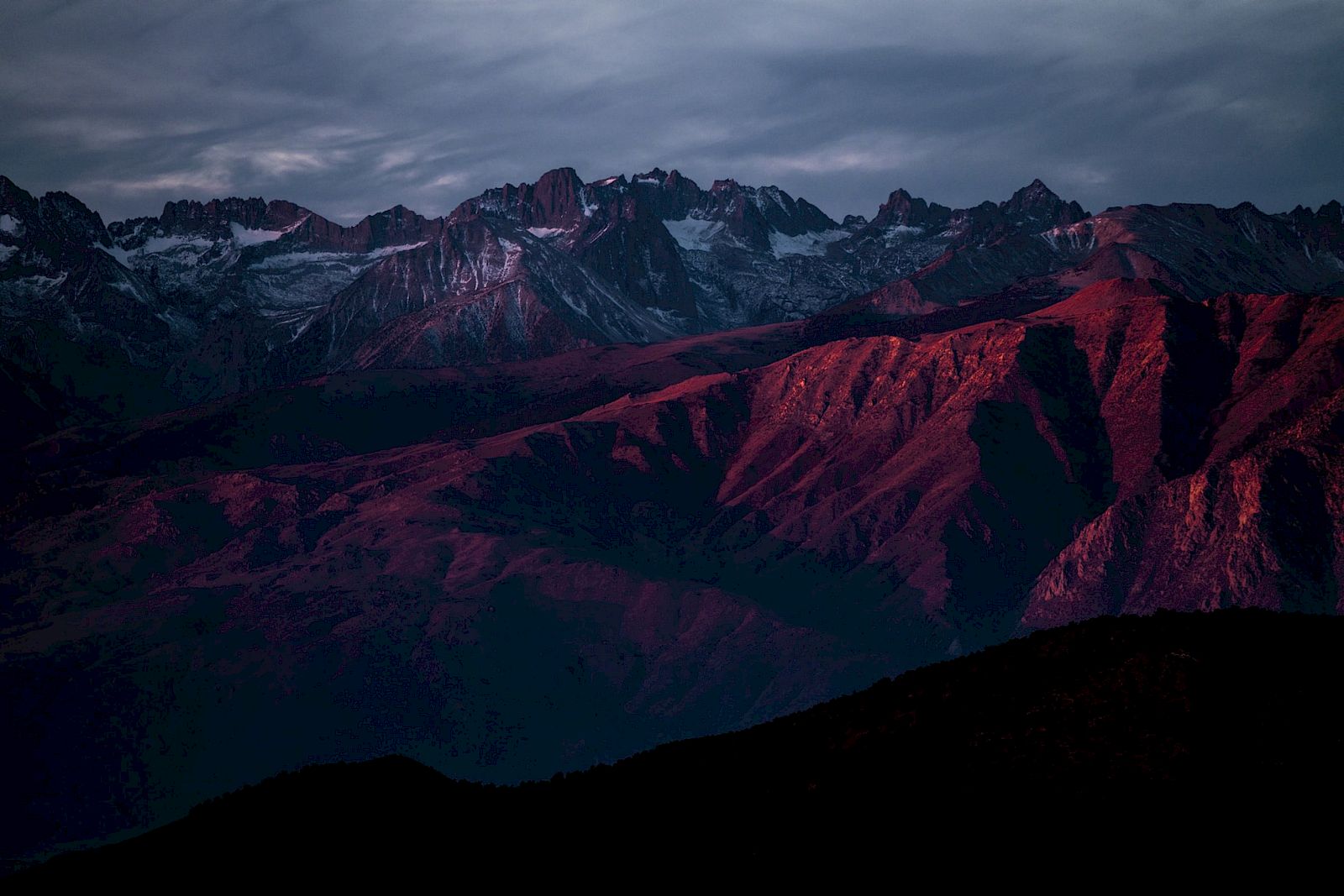 A mountain range at dusk with dramatic shadows and a red hue on rocky peaks under a cloudy sky.