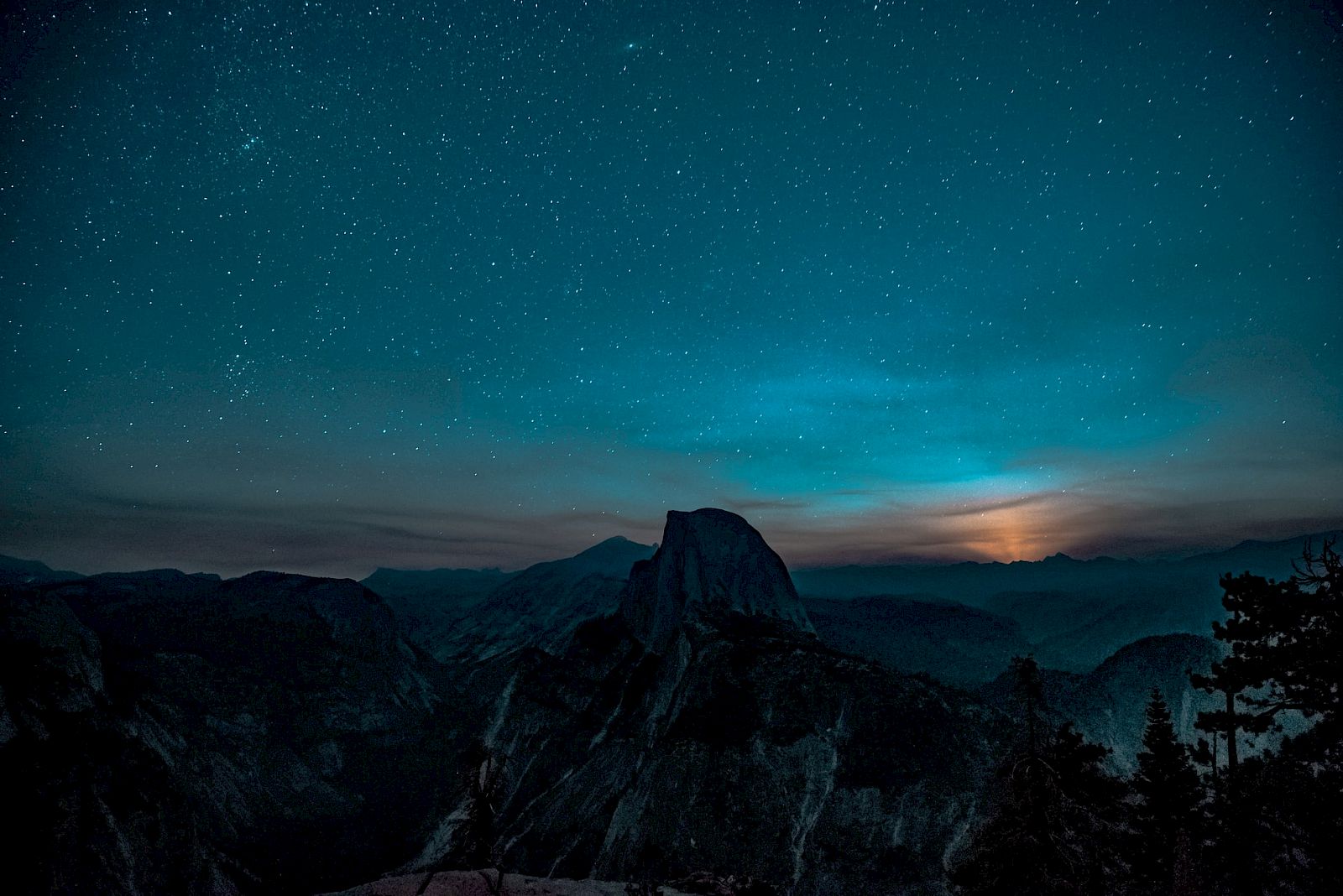 A starry night sky over a mountainous landscape with silhouetted trees and a faint glow on the horizon, creating a serene scene.
