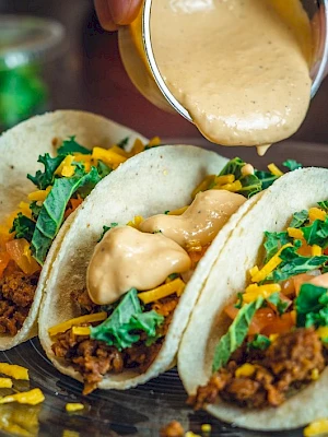 Three tacos filled with ground meat, shredded cheese, tomatoes, and greens, topped with a creamy sauce being poured from above.