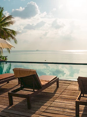 Poolside scene with lounge chairs, umbrellas, and palm trees beside a calm sea under a cloudy sky, creating a relaxing atmosphere.