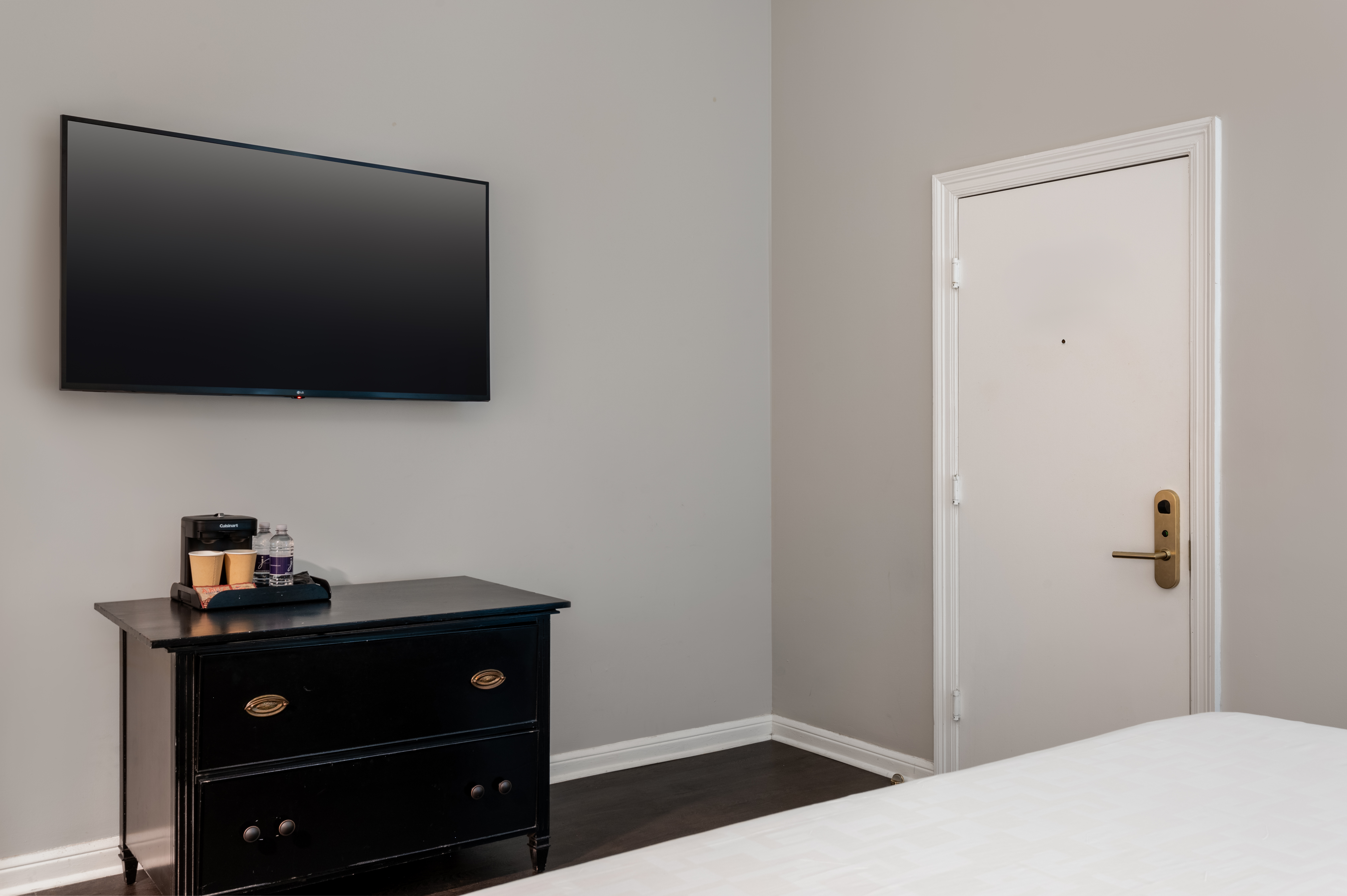 A hotel room with a TV mounted on the wall, a black dresser with a coffee maker and cups, and a bed with white sheets.