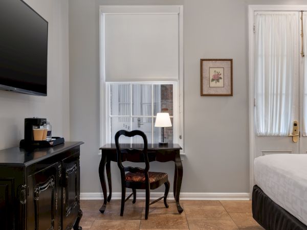 A room features a bed, TV, ornate desk with chair, a lamp, coffee maker, and framed art, with soft lighting and neutral decor.