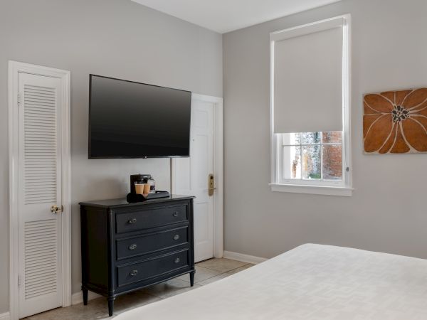 A bedroom with a TV, dresser, and coffee maker. Artwork on the wall and a window with a blind complete the decor.