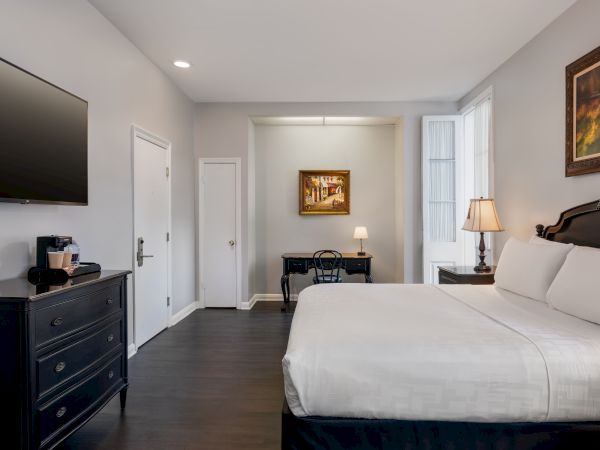 A modern hotel room with a large bed, wall-mounted TV, desk, lamps, and artwork, featuring neutral tones and minimalistic decor.