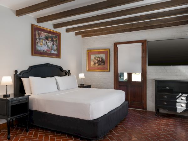 A cozy bedroom with white walls, featuring a black-framed bed, bedside lamps, paintings, wooden beams, a TV, and brick flooring.