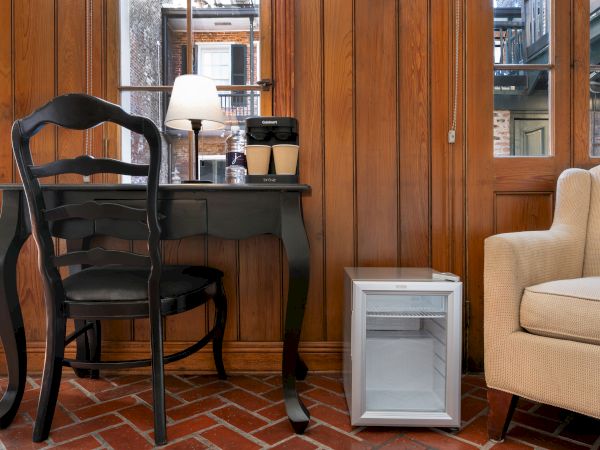 The image shows a black desk with a chair, a table lamp, coffee cups, a beige armchair, and a small fridge on a brick floor.
