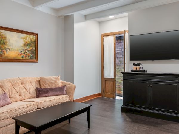 A living room with a beige sofa, black coffee table, wall-mounted TV, wooden cabinet, painting, and an open door.
