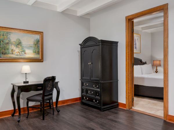 A room with a small black desk, chair, lamp, black armoire, painting, wood trim, and an open door to a bedroom with a bed and lamp.