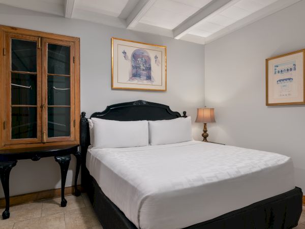 The image shows a cozy bedroom with a double bed, two artworks on the walls, a wooden cabinet, and a lamp on a small table.