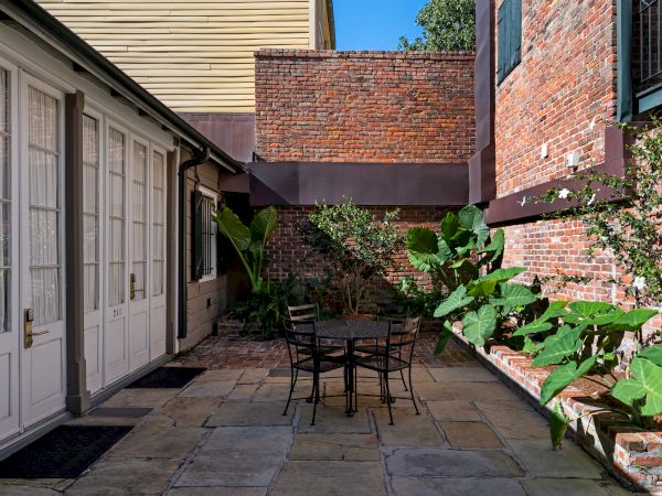 A cozy patio area with a metal table and chairs, surrounded by brick walls and lush greenery, under a clear blue sky.