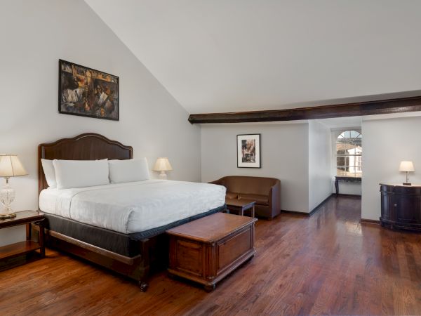 A spacious bedroom with a large bed, wooden floors, bedside tables with lamps, artwork on the walls, and natural light shining through.