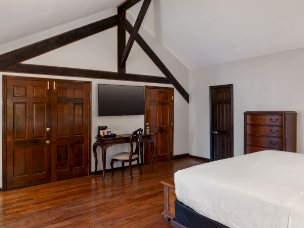 A bedroom with wooden floors, a bed, a dresser, a TV on the wall, a small desk with a chair, and exposed wooden beams.