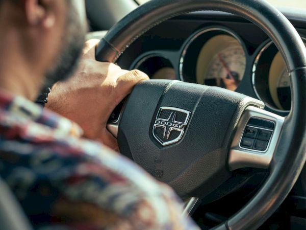 A person is driving a car, holding the steering wheel with a logo visible, focused on the road ahead.