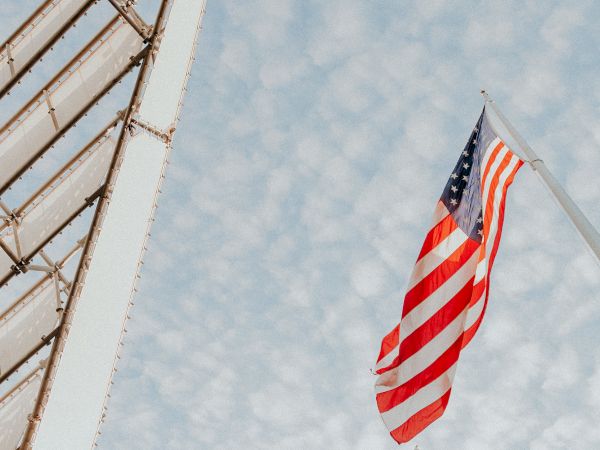 An American flag is waving on a pole against a cloudy sky, with part of a building structure visible on the left side.