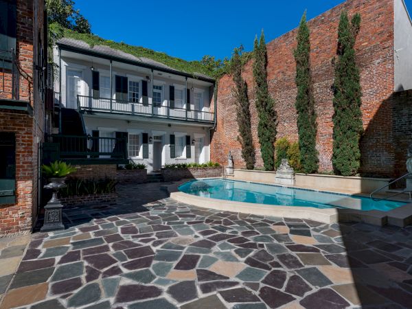 The image shows a courtyard with a small pool, surrounded by brick buildings and a stone-tiled floor, featuring tall trees and statues.