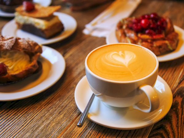 A cappuccino with latte art on a wooden table, surrounded by pastries on plates, including one topped with cherries.