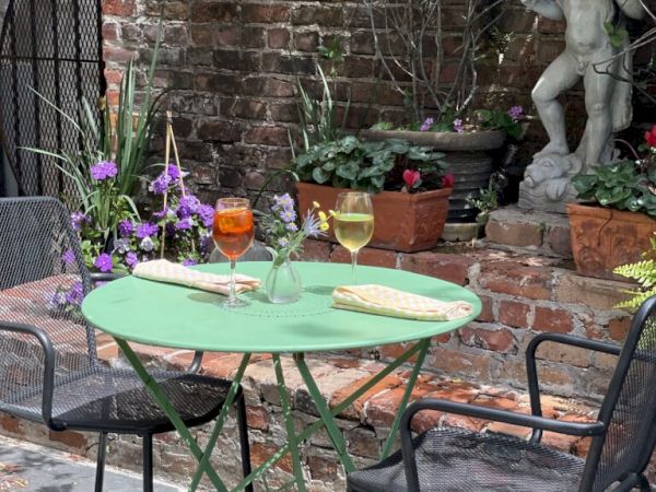 A small garden patio with a green table, two drinks, and a baguette. There's a statue in the background among potted plants.