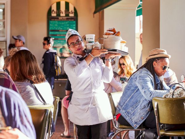 A waiter carries a tray in a busy cafe with seated patrons around, under warm lighting and green awnings.