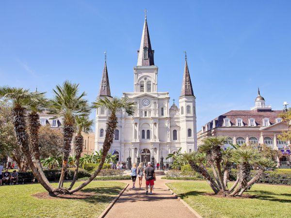 The image shows a historic building with three spires, surrounded by palm trees and a walking path on a clear sunny day.