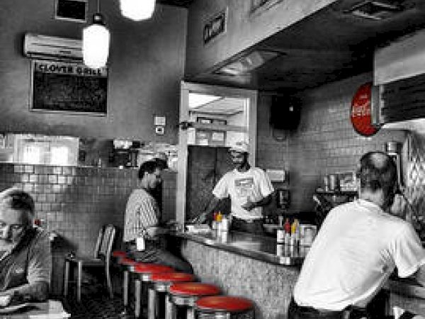 A vintage diner with red stools, black-and-white decor, patrons sitting at the counter, and a server preparing orders in a cozy atmosphere.