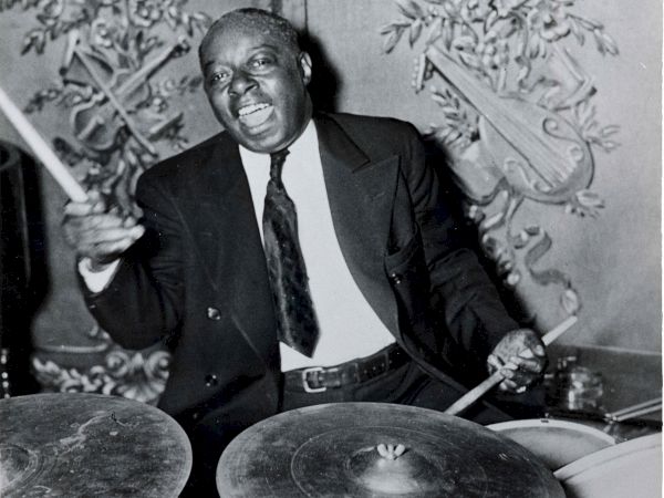 A person in a suit is energetically playing the drums, surrounded by ornate wall decor.