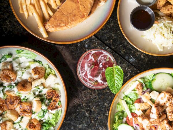 An assortment of dishes including fries with a sandwich, salads with chicken and scallops, and a drink garnished with mint leaves.
