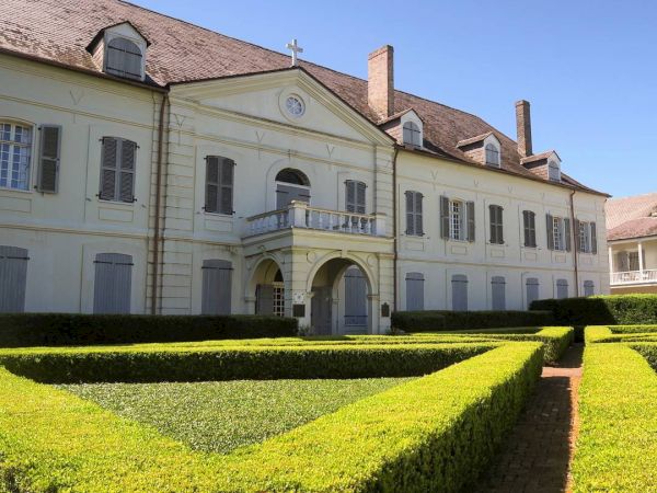 A large, classic villa with a symmetrical façade and arched entrance is surrounded by neatly trimmed hedges under a clear blue sky.